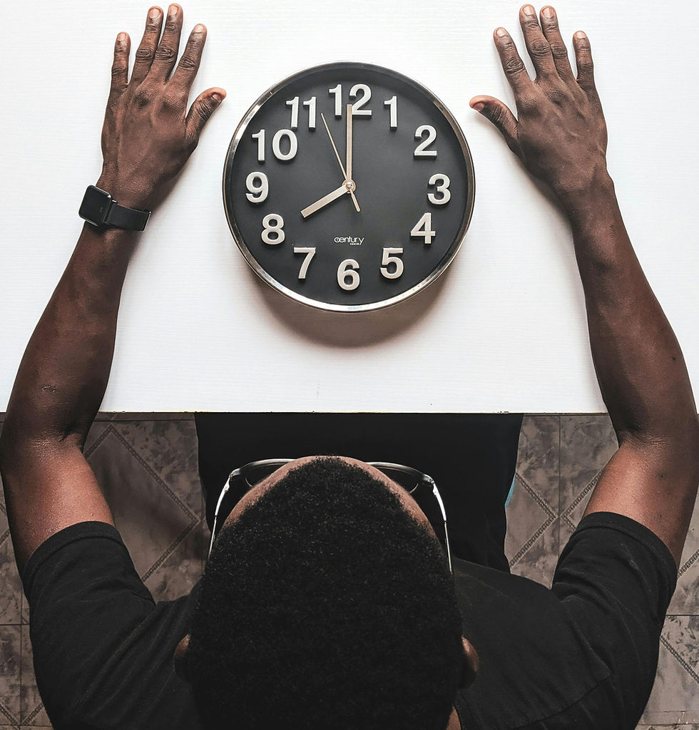Person with outstretched arms facing a black wall clock on white table, reading 10:10. Wearing a black shirt and watch. Gray tiled floor.