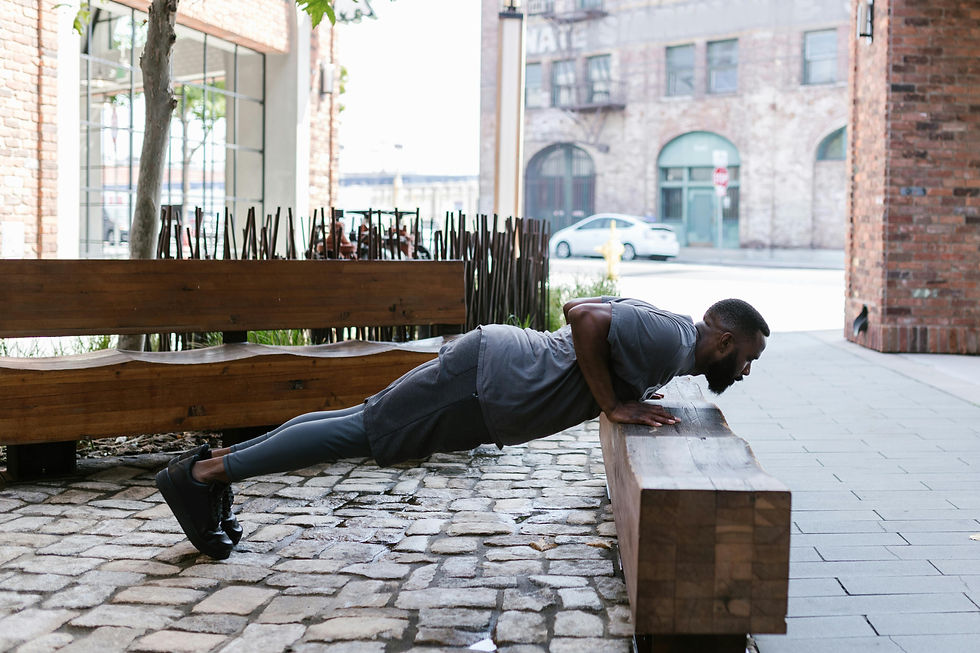 Man doing push-ups on a wooden bench in an urban setting. He wears gray workout clothes. Brick buildings and a parked car are in the background.