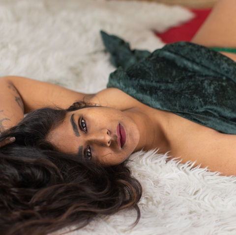 Woman with light-brown skin covered in a green suede blanket laying on a bed with her dark brown hair strewn about