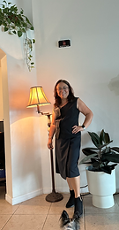 A picture of the CEO of the company standing by a lamp and by a plant, with her hand by the hips, wearing a black dress