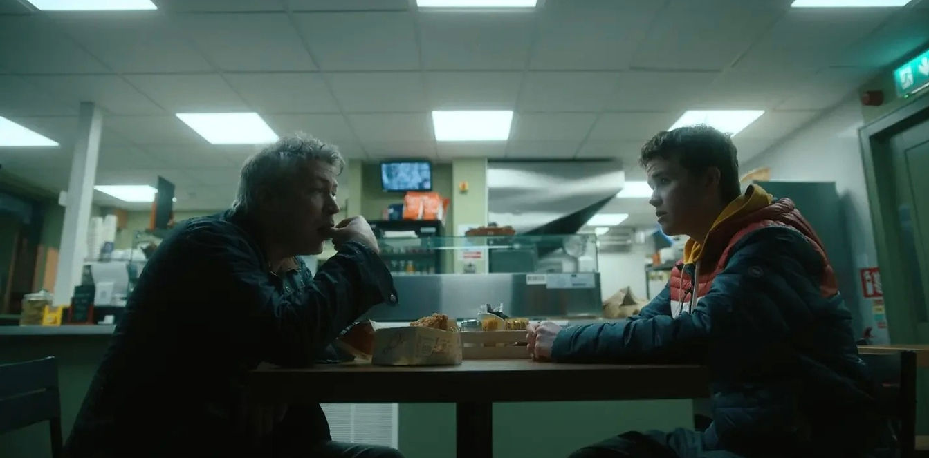 Two men sit at a table, talking in a dimly lit restaurant setting.