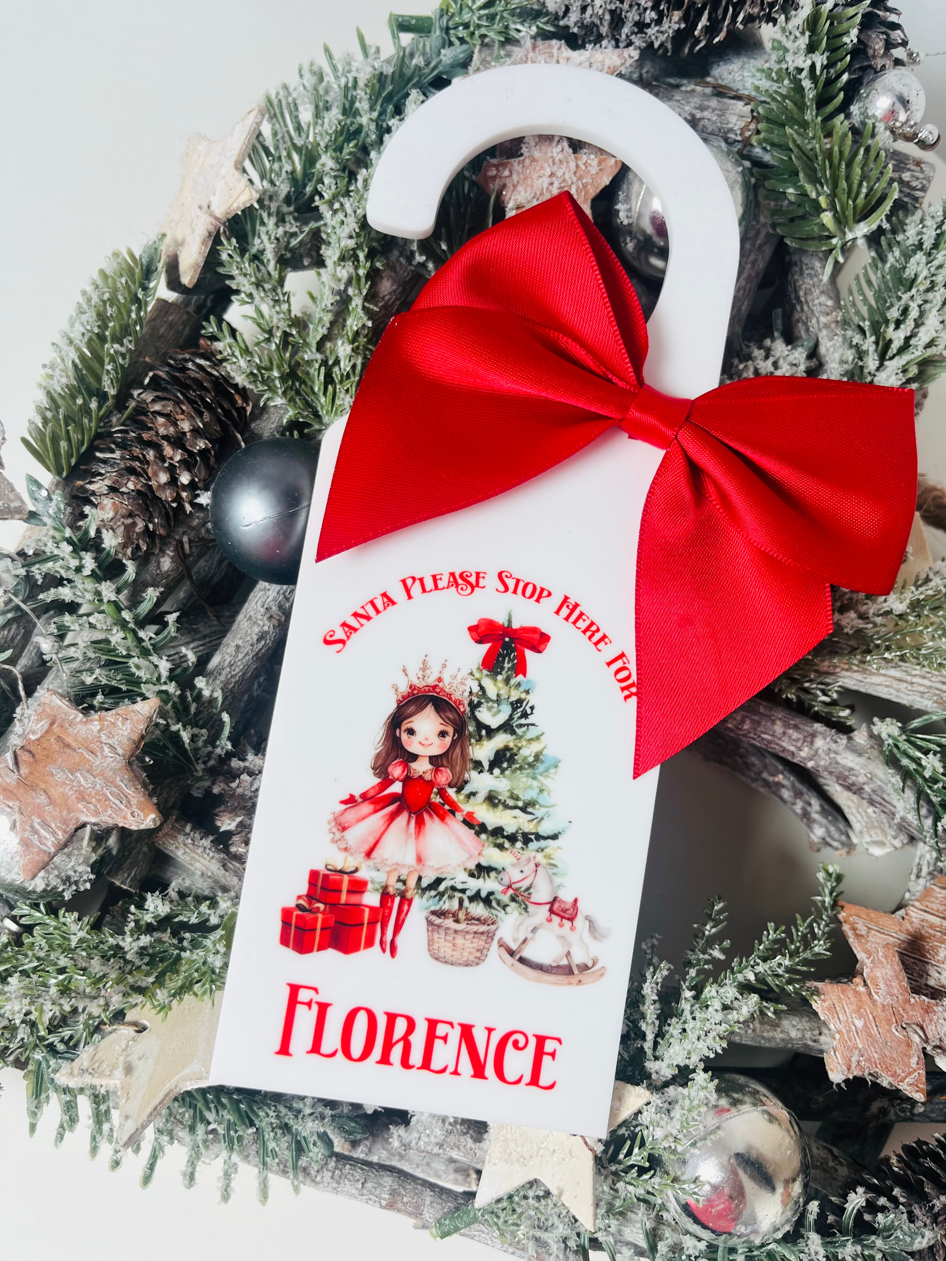 A white acrylic door hanger with a big red satin bow. The image is a red sugarplum fairy and christmas tree. It says "Santa, please stop here for 'name'"