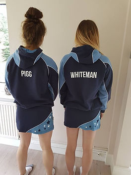 Two girls standing next to each other, wearing the St Neots Netbal match dress and the netball hoodie, facing backwards.