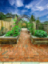 A symmetrical view of a small courtyard vegetable garden featuring rustic oak raised beds and a pathway made from reclaimed clay cottage pavers. Overhead, natural willow arches support climbing beans with vibrant green leaves and orange-red blossoms. The design balances beauty and practicality, enclosed by wooden fencing with a pale garden shed at the rear, all set within a charming and luxurious residential setting. Small garden design and landscaping by luxury gardens.