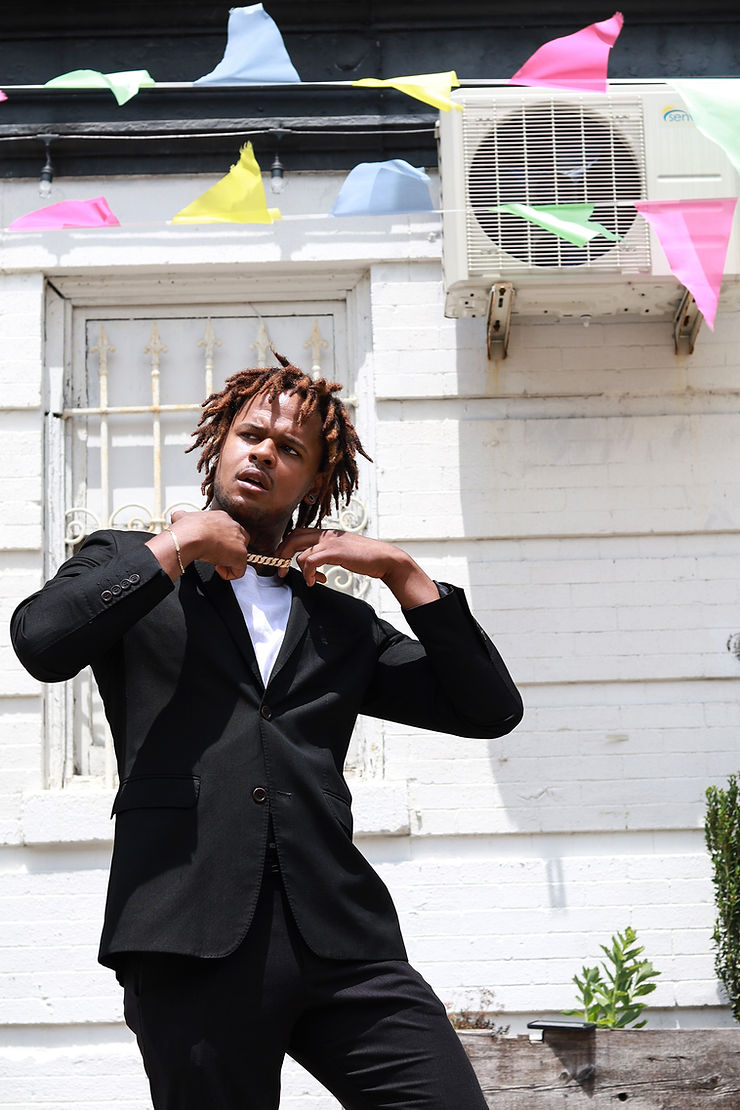 Jamaal Singleton in black suit adjusts chain, standing in front of a white brick wall with colorful flags. Bright, sunny setting.