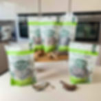Five Forthglade Natural Cuts pet treat bags on a kitchen counter with dried fish and meat, set against modern white cabinets.