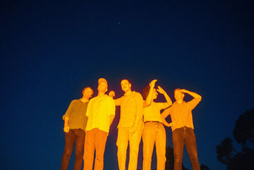 Some songs feel like postcards. Others feel like the walk you took before you ever thought to write one. Rolling Blackouts Coastal Fever deal in the latter—motion, light, the hum of a city passing through you before you’ve had time to name it.
Out of Melbourne, the band built their sound on interlocking guitars that don’t so much compete as converse. Three voices, three lines, each one threading through the others with an almost absent-minded precision. There’s a looseness to it, but not carelessness. Their music moves the way people move through familiar streets—quickly, instinctively, guided by something half-conscious.
They’ve often been described as jangly, bright, even breezy, but that misses the undercurrent. Beneath the propulsion, there’s a restlessness, a sense that all this movement is happening for a reason that remains just out of frame. Their songs travel well, but they’re rarely about arrival.
Sunburned in London carries that feeling with particular clarity. It opens like a day already in progress, no grand introduction, just the continuation of something you’ve stepped into midway. The guitars shimmer without settling, each one catching the light at a slightly different angle. The rhythm pushes forward, steady but not urgent, like footsteps that know where they’re going even if you don’t.
The title itself suggests a small dislocation—a body out of place, marked by a different climate, a different sky. London, with its grey reputation, becomes the backdrop for something faintly absurd: sunburn as evidence of elsewhere, of having carried another world with you. It’s a quiet kind of contradiction, and the song leans into it without explanation.
The vocal drifts through images rather than declarations. Nothing is pinned down for long. Streets, sensations, passing impressions—they arrive, they dissolve. There’s a feeling of being both present and slightly removed, as though the narrator is watching their own experience unfold from a short distance away.
What makes the track linger is its refusal to dramatize that distance. There’s no crisis here, no sharp turn inward. Just the steady accumulation of detail, the sense of a day stretching on, shaped by small, almost imperceptible shifts. It captures that peculiar state of travel where everything feels meaningful and disposable at once.
Rolling Blackouts Coastal Fever have a gift for this kind of balance. They let the music carry you forward while the lyrics quietly unsettle your footing. You move, you listen, you piece things together, and still something remains unresolved.
Sunburned in London doesn’t offer a revelation. It offers a condition. A body in motion, slightly out of sync with its surroundings, catching light where it shouldn’t, holding onto it a little longer than expected. And in that small, lingering warmth, you find something like recognition.