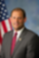 Portrait of a smiling man in a gray suit and red tie, posed before a U.S. flag and blue backdrop.