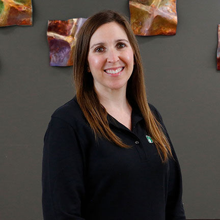 Sarah Prechtel: She is a white woman with long, straight brown hair. She is smiling at the camera with a black polo shirt with DEAF, Inc. Logo. The background is grey with decorative squares.