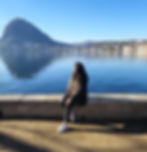 uma mulher sentada numa mureta olhando para o horizonte com montanhas, um lago e casas.