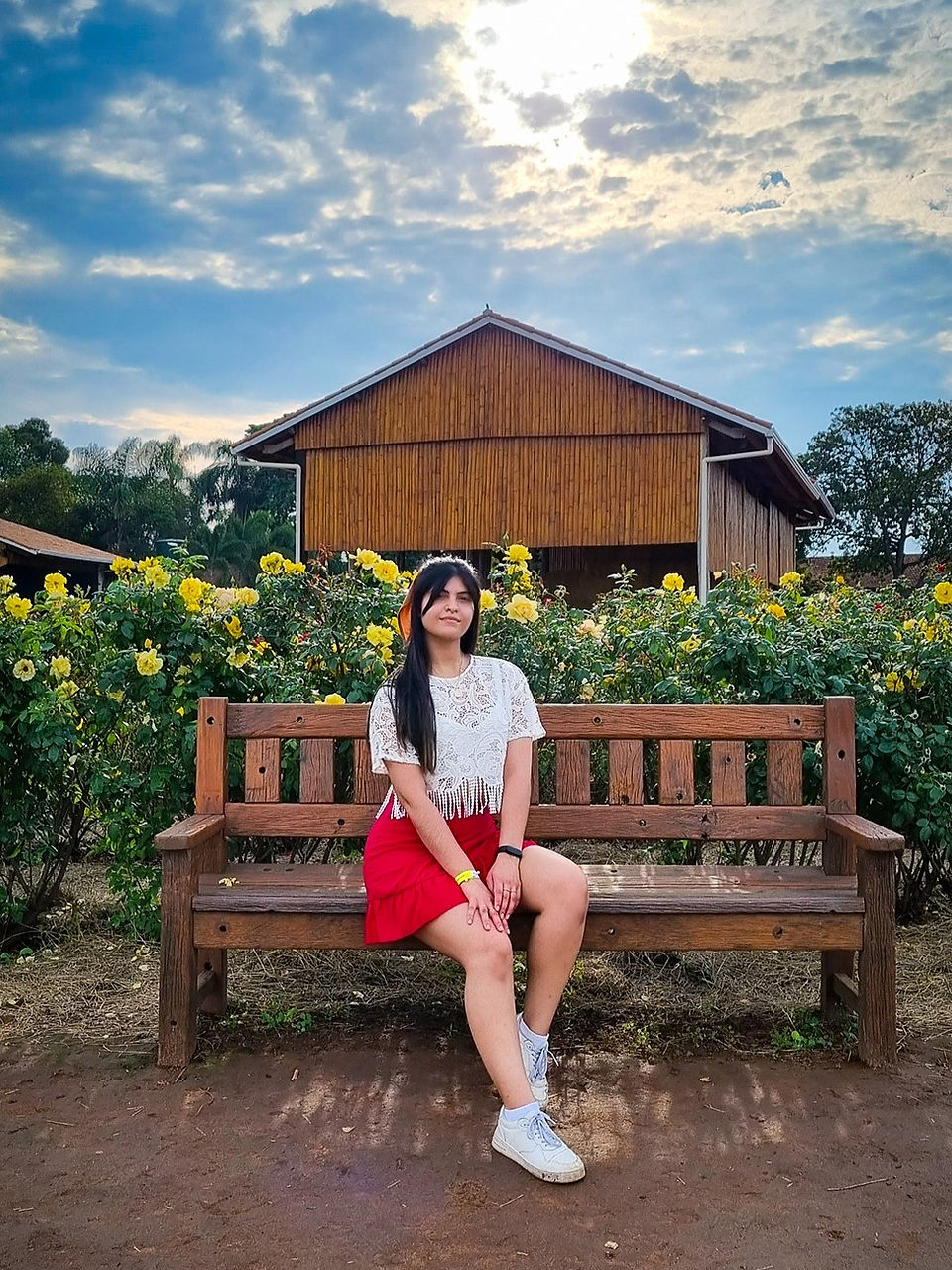 Uma menina sentada em um banco de madeira no meio de um jardim com uma casa ao fundo