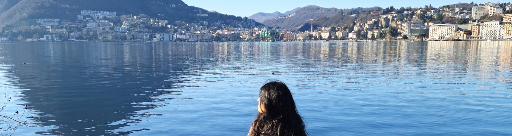 Uma pessoa sentada em uma mureta, olhando para o horizonte onde contém um lago e montanhas ao fundo.