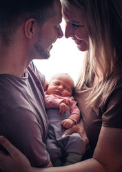 Newborn-/Babyfotografie Braunschweig TINA KERCHER WEDDING Eltern mit Baby in der Mitte. Sie stehen sich gegenüber, seitlich zur Fotografin, das Baby liegt inmitten beider in deren Armen. Die Eltern sehen sich glücklich an. Sie stehen im Gegenlicht, 