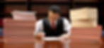 Man in a suit writing at a desk, surrounded by stacks of red-covered books. Wooden bookshelf in the background. Focused mood.