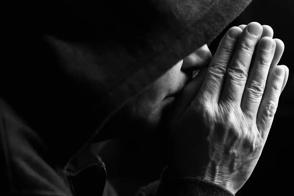 Close-up of a person in a hood, hands pressed together near face, conveying contemplation. Black and white, dark shadows enhance mood.