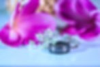 This image appears to depict wedding rings and accessories. The main focus of the image is a pair of diamond engagement and wedding rings resting on a dark surface, surrounded by a vibrant pink flower petal. The background is blurred, but appears to be a light blue or aqua color. The overall composition and styling suggests this is an image related to a wedding or engagement celebration.