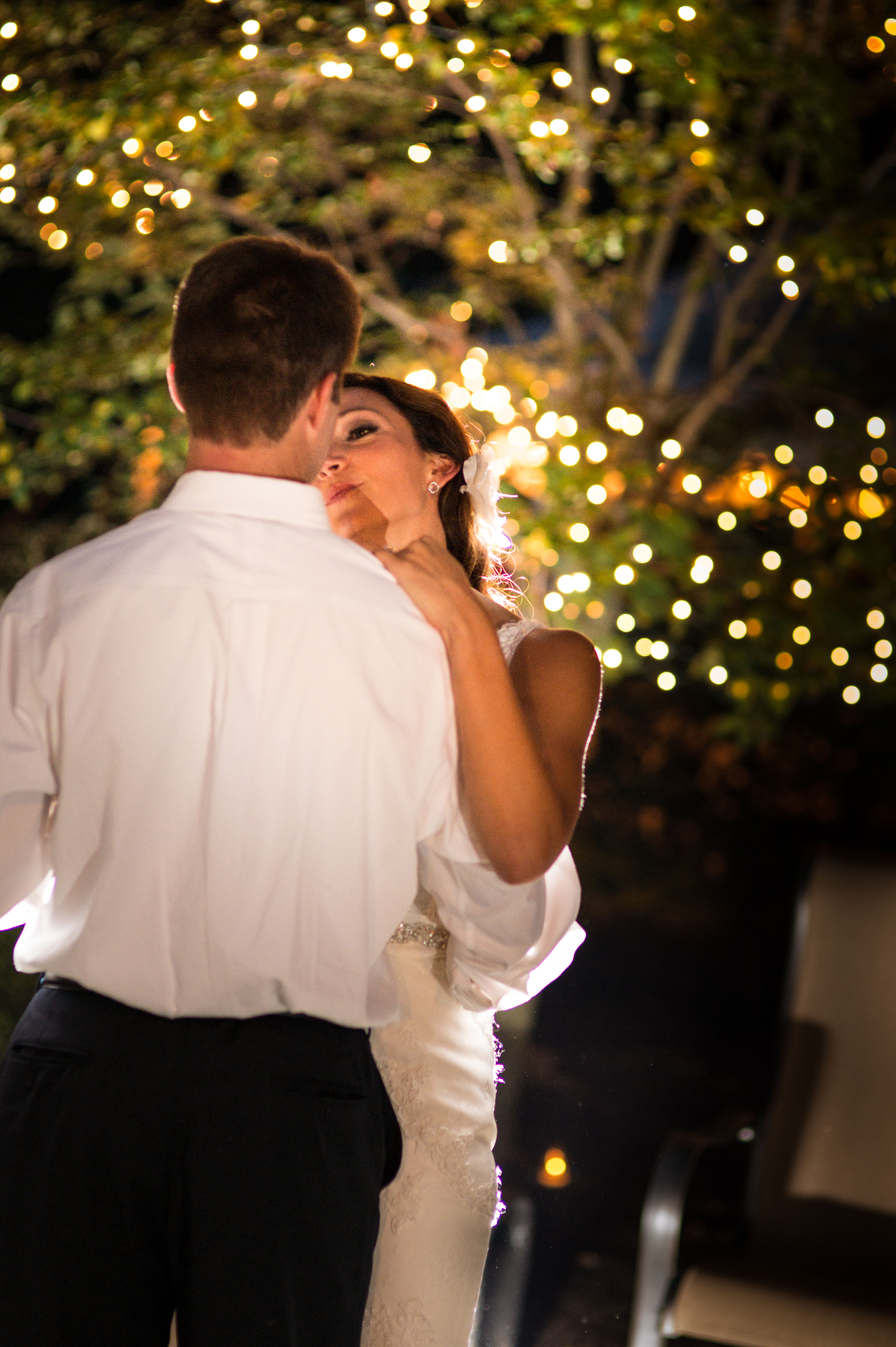 A bride and groom are standing close together, facing away from the camera towards a backdrop of twinkling lights. The groom is wearing a white shirt, and the bride is in a white wedding dress with her hair up. They appear to be having an intimate moment. There is a warm glow around them, suggesting it might be an evening event.