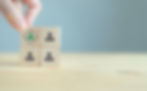 Hand arranging wooden blocks with icons of people. One block shows a green figure under a crosshair, against a plain background.