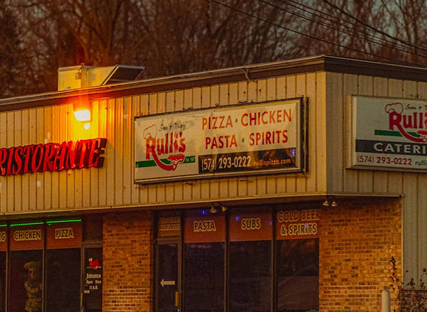 Rulli’s Italian Restaurant on County Road 17 in Elkhart, Indiana, with the storefront and signage for pizza, pasta, chicken, and spirits visible at dusk.