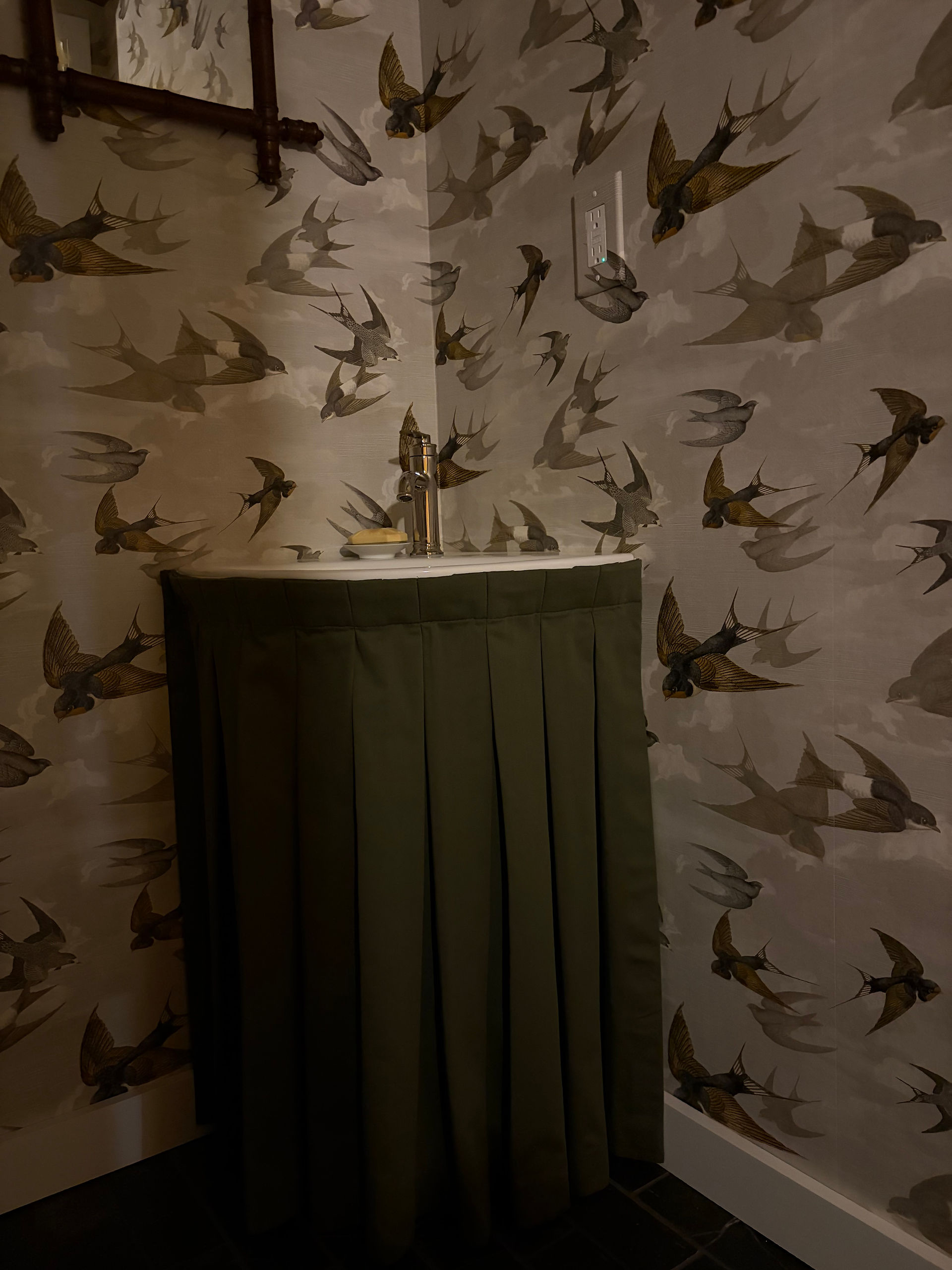 Basement powder bath renovation featuring swallow wallpaper and vintage mirror with skirted corner pedestal sink