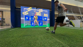 Pessoas se divertindo no Chute ao Gol interativo da PixelSAV no Shopping Mueller, com tela LED grande e goleiro virtual