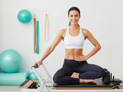 Imagem horizontal realista em 4:3 de uma mulher de 35 anos sorrindo confiante realizando exercício de Pilates no reformer, com foco na região pélvica, em estúdio moderno e luminoso. Paleta de verde-água, branco e bege transmitindo força, saúde e bem-estar feminino.