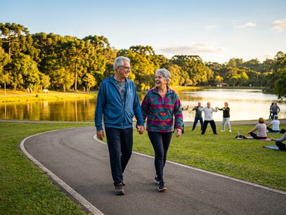 Casal de idosos caminhando juntos em parque ensolarado com boa postura e expressão alegre, representando envelhecimento ativo, condicionamento físico e qualidade de vida na terceira idade.