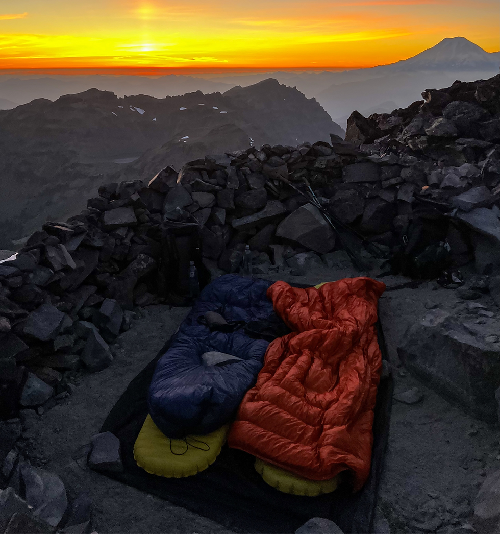 Two sleeping bags, blue and orange, on a rocky mountain peak at sunrise. The sky is orange, distant mountains visible, creating a serene mood.