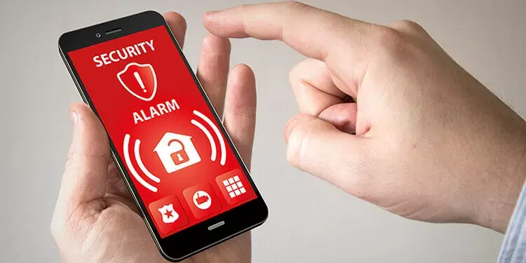 Hand holding a smartphone with a red security alarm screen showing home protection features and alert icons.