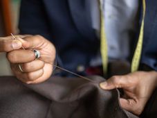 Tailor's hands stitching brown fabric with needle and thread, wearing a metal thimble on one finger. A tape measure is visible around the sewer's neck, blurred in the background.
