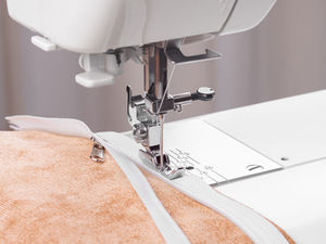 Close up of a sewing machine stitching a white zipper onto tan fabric. 