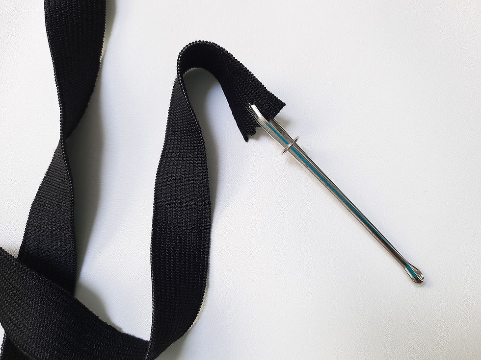 A metal clip style bodkin and a strip of black elastic on a white background. The bodkin is clipped to the end of the elastic, with a sliding ring locking the bodkin closed.