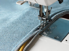 Close up of sewing machine stitching piping on heavy blue fabric. The fabric partially encloses a yellow cord.