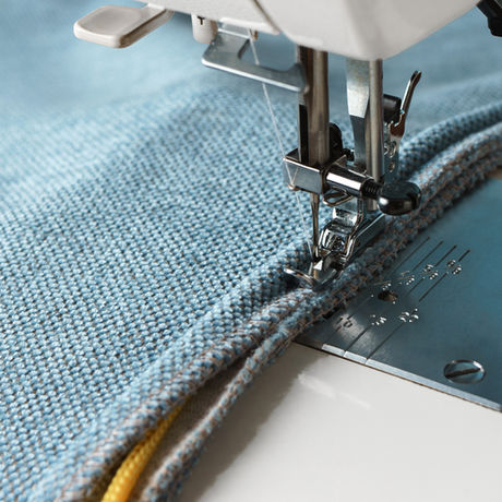 Close up of sewing machine stitching piping on heavy blue fabric. The fabric partially encloses a yellow cord.