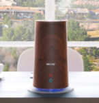 Wood-patterned humidifier emits mist on a white table in a room with large windows. Trees and buildings are visible outside.