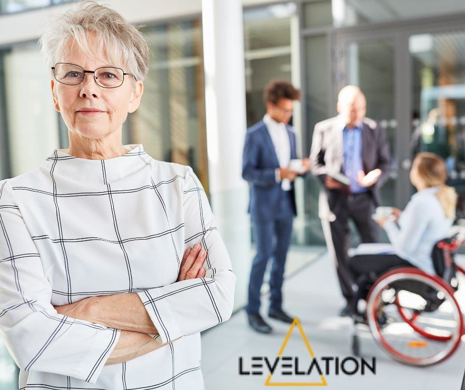 Empowering Women In Tech - A white haired white lady wearing glasses and a white bold checked dress in the foreground with her arms folded, she looks authoritative, blurred in the background are two men in suits and a lady in trousers and a blouse in a wheelchair against a business office building