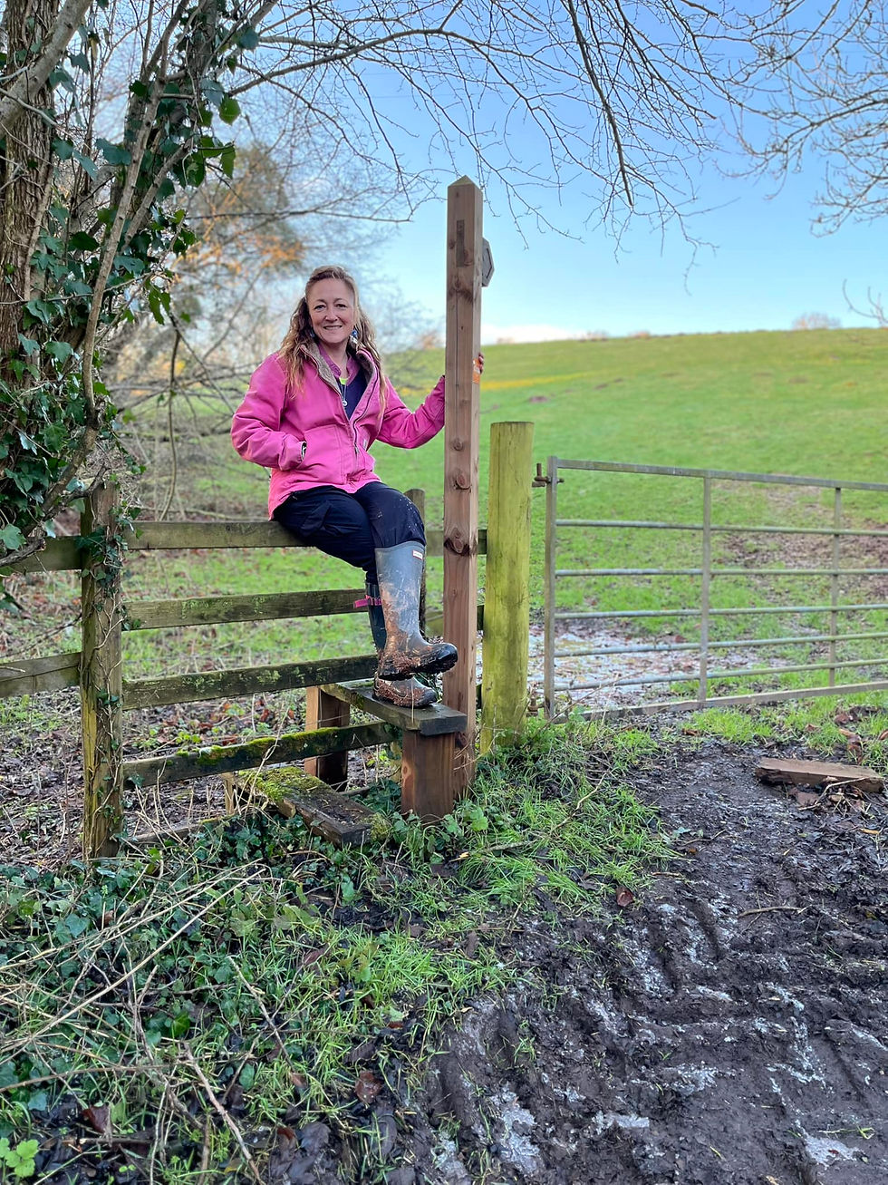 World Wildlife Day - Miriam dressed in a pink coat, trousers and wellies is sitting on a style next to a field and fence under a tree