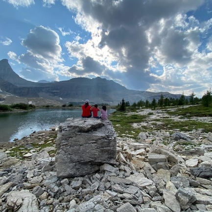 A mom and her two children sitting on a boulder looking out over a high alpine lake with mountains in the background