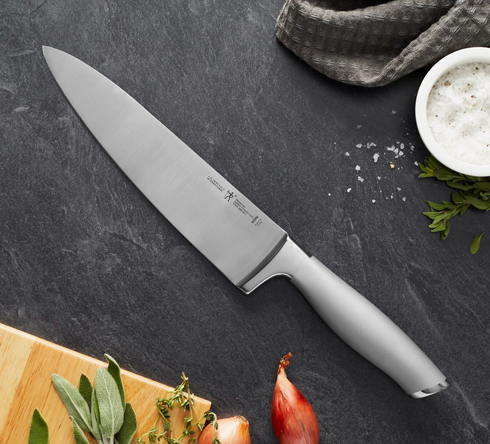 Chef’s knife on dark stone countertop with salt bowl, herbs, shallot, and cutting board; clean, moody kitchen still life