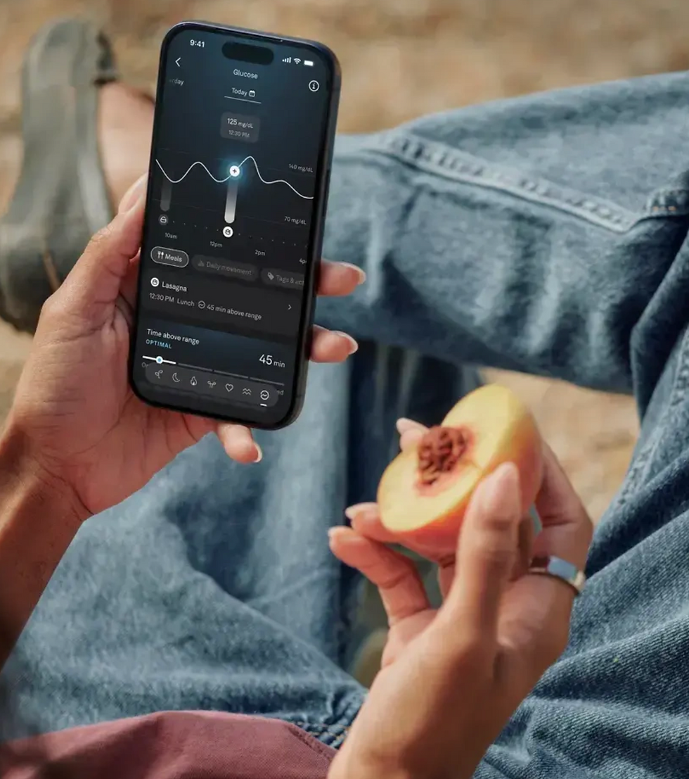 Person holds a smartphone showing a glucose graph while eating a peach, with jeans-clad legs in the background.