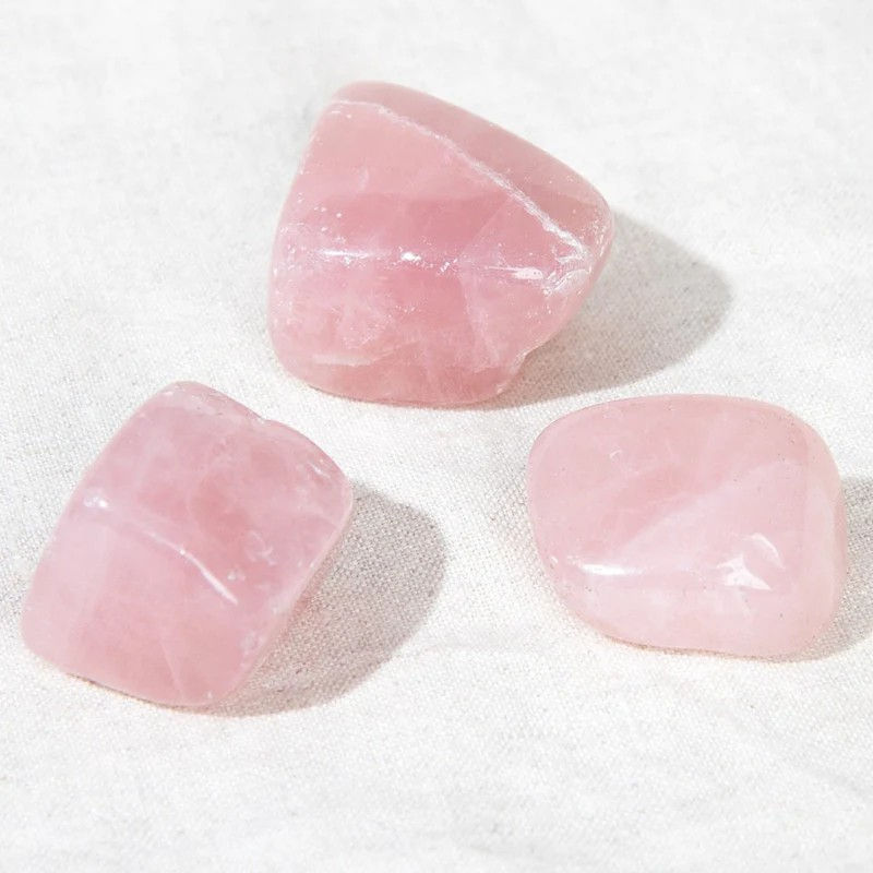 Three polished pink rose quartz stones on a white fabric background, exhibiting smooth surfaces and soft pastel hues.