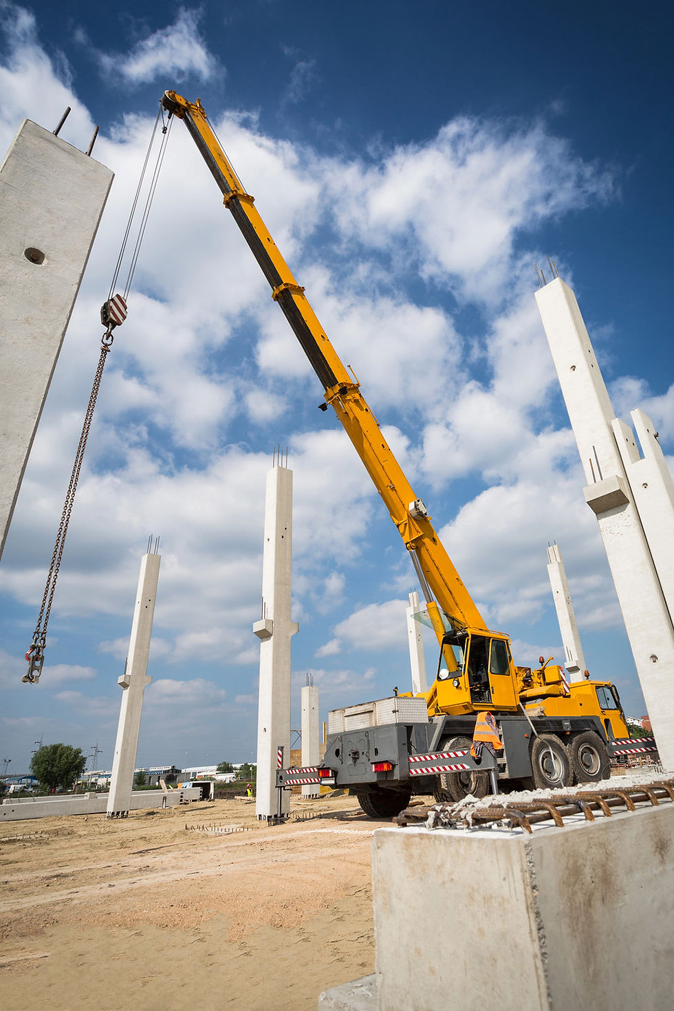 A large yellow mobile crane on a construction site, lifting a concrete beam against a blue sky.