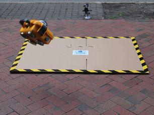 A yellow HoistCam camera in mid-air, about to impact a piece of cardboard on a brick sidewalk.