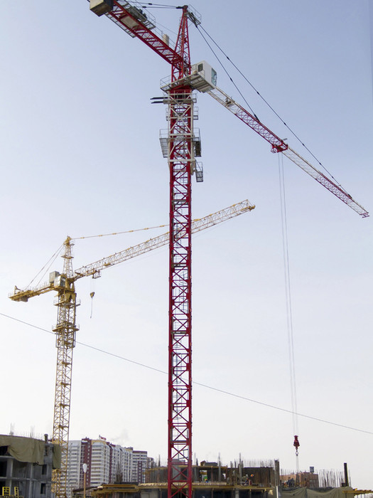 Two tower cranes at a construction site, one painted red and white in the foreground and another yellow in the background. Both cranes are actively positioned above a multi-story building under construction, with workers and scaffolding visible on the site below.