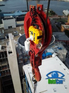 A red crane hook with a yellow HoistCam attached, and a tablet displaying the camera's view of a construction site.