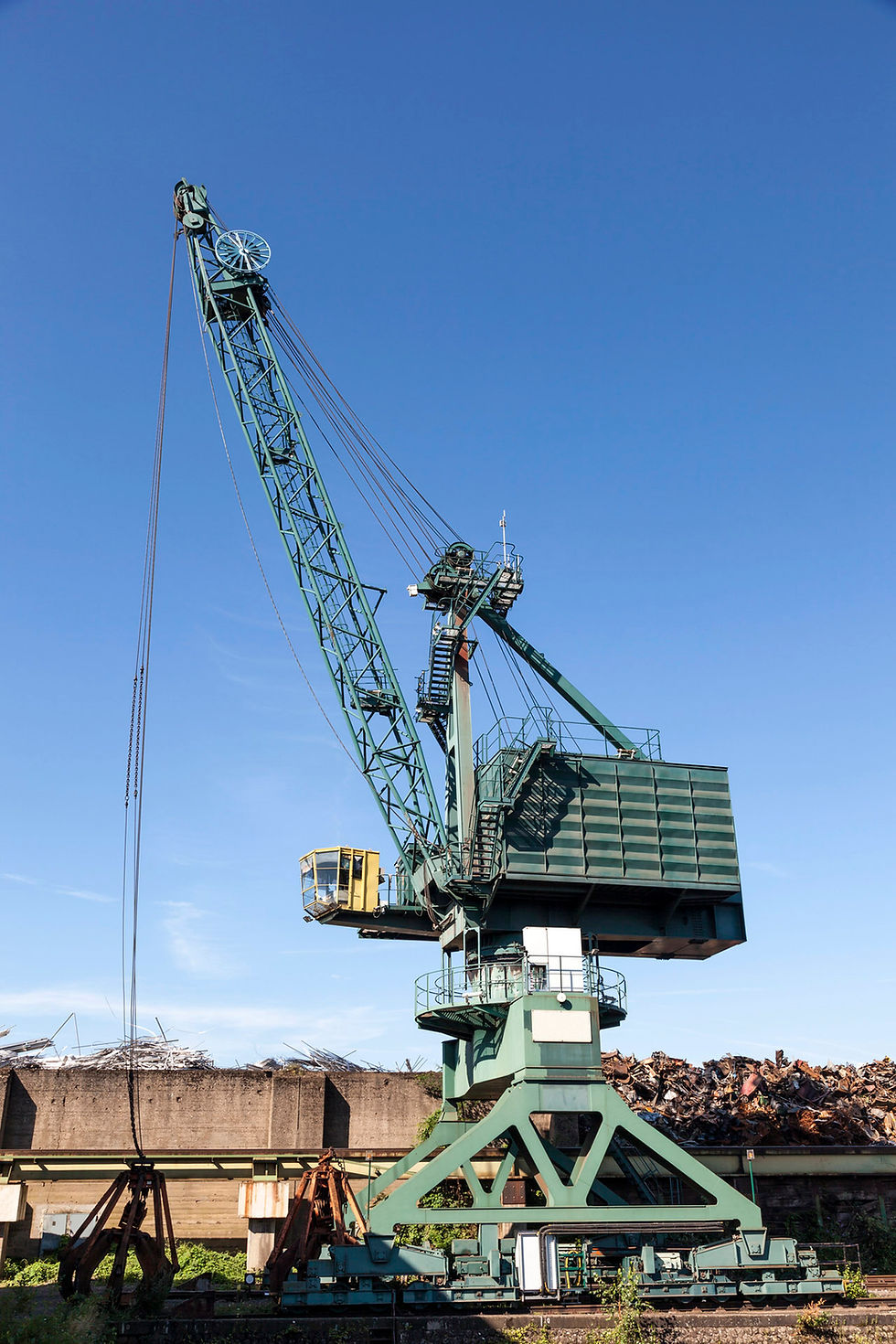 A large green and yellow grapple crane stands against a clear blue sky, next to a pile of scrap metal.