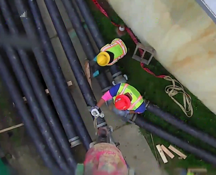 Two workers guide a crane hook to lift large industrial pipes at a construction site.