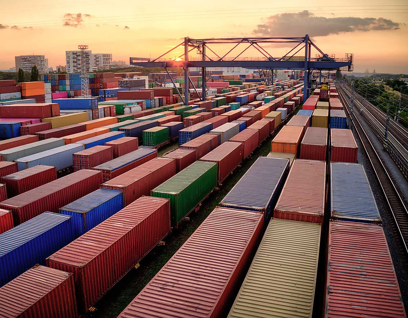 Intermodal yard filled with colorful shipping containers at sunset, with rail tracks and gantry cranes in the background.