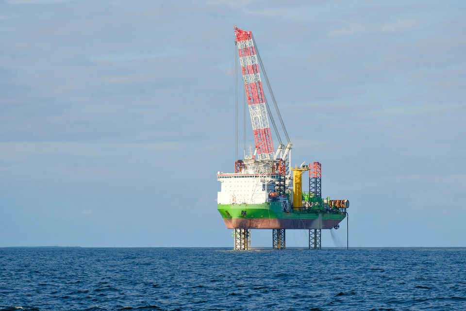 An offshore jack-up vessel equipped with a large red and white crane operates in open water under a blue sky. The vessel is supported by extended legs anchored to the seabed, and is used for heavy lifting and marine construction, such as wind turbine or oil platform installation.