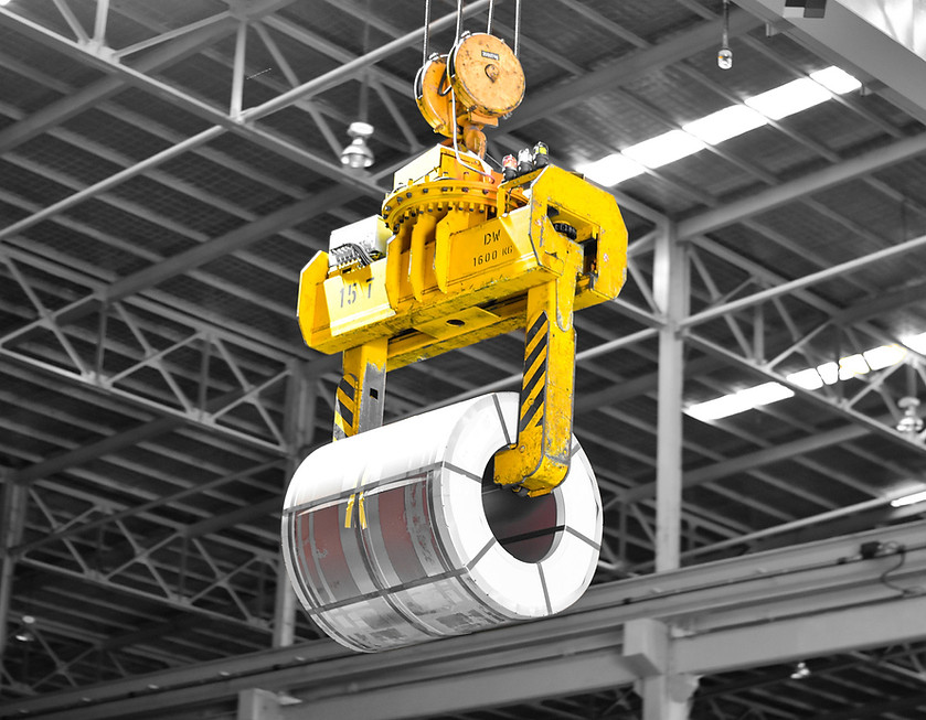 Overhead crane lifting a large metal coil with a yellow coil grab inside an industrial facility.